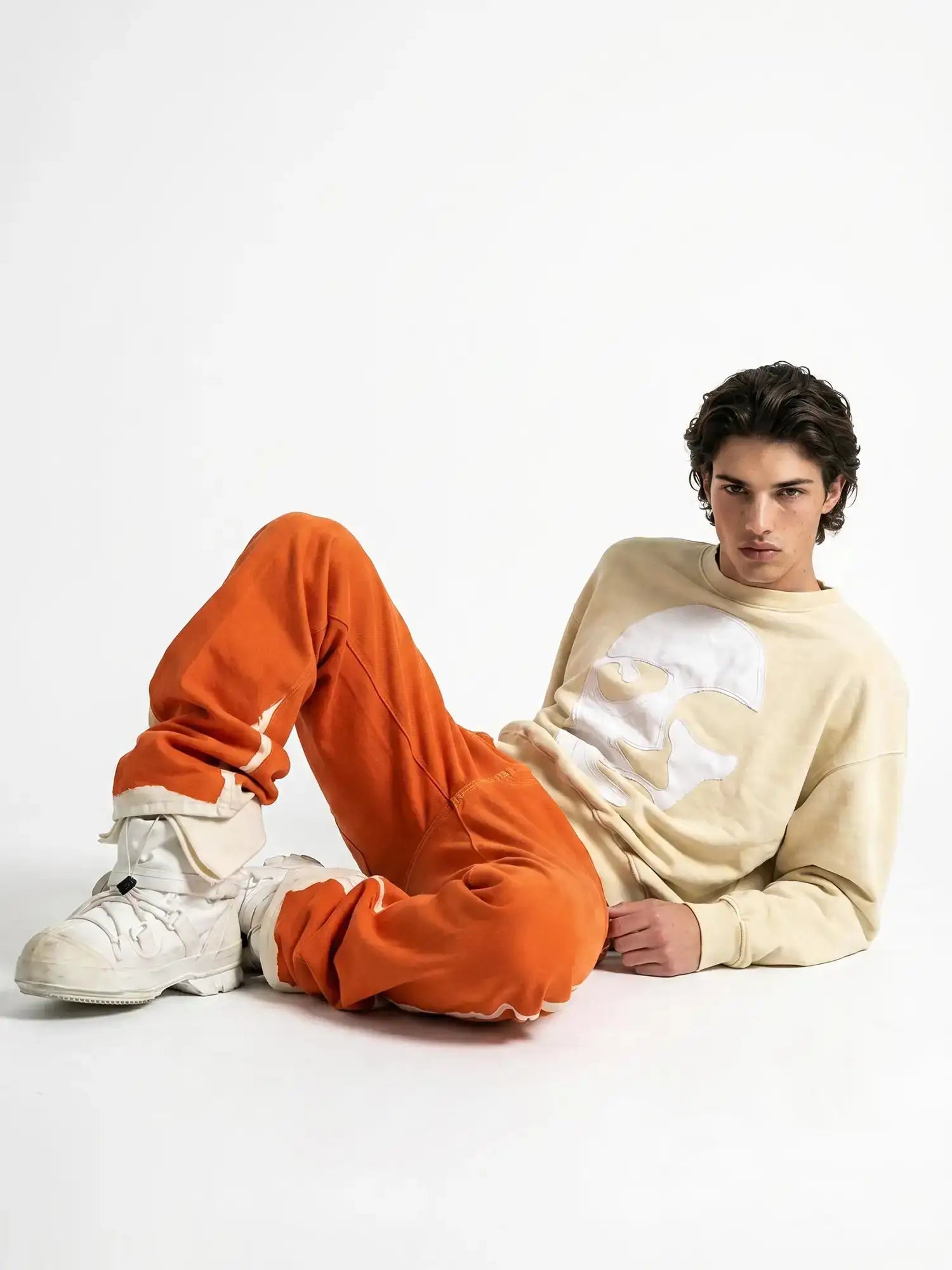 Male model wearing beige face-appliqué sweatshirt paired with orange trim pants