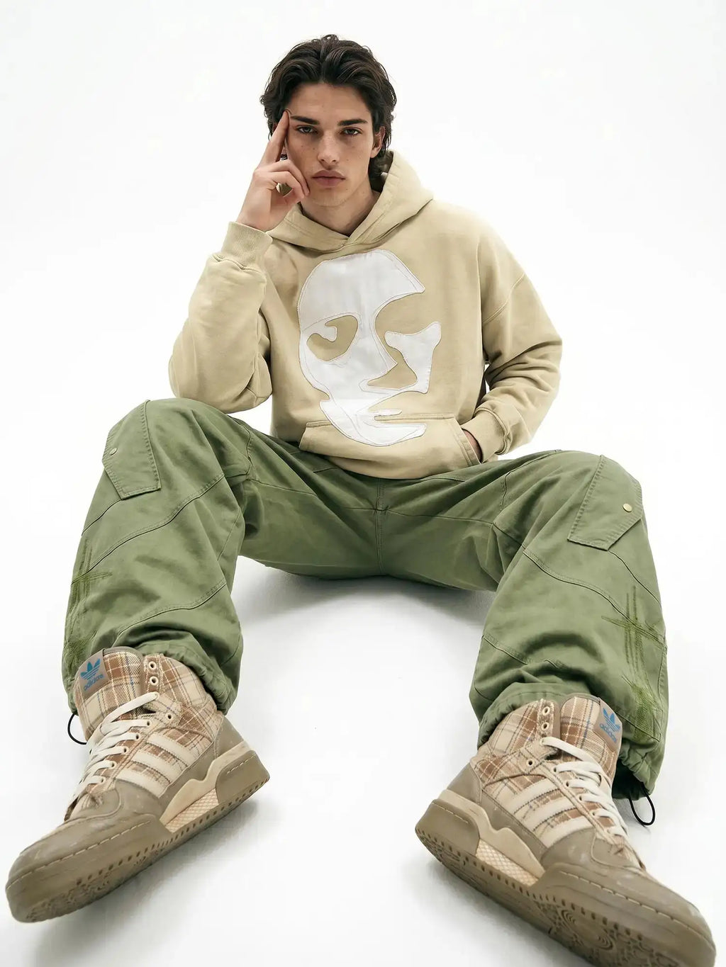 Beige hooded sweatshirt with white face design styled with olive cargo pants and plaid sneakers