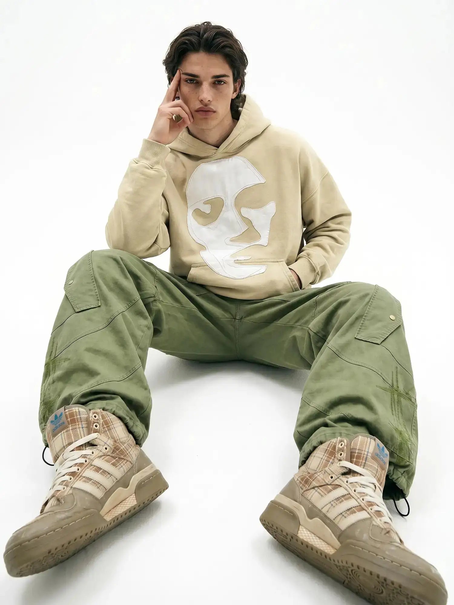 Beige hooded sweatshirt with white face design styled with olive cargo pants and plaid sneakers